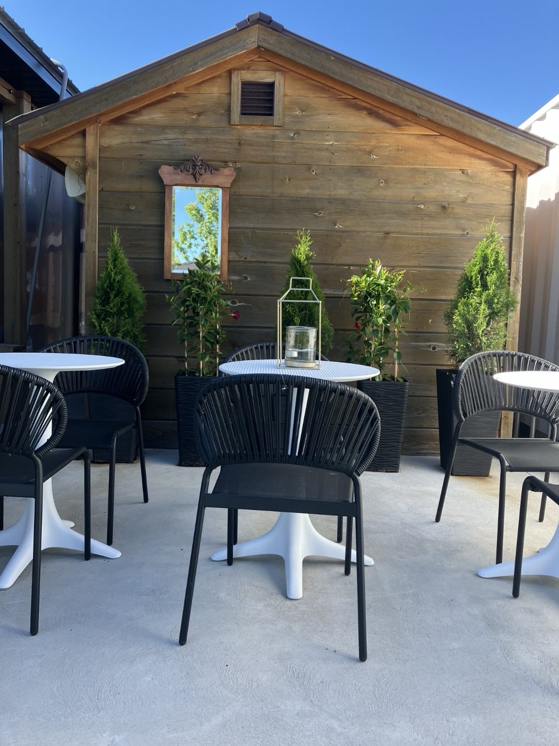 Bistro 93 patio with black rope chairs, white tables and potted greenery against a weathered-wood wall